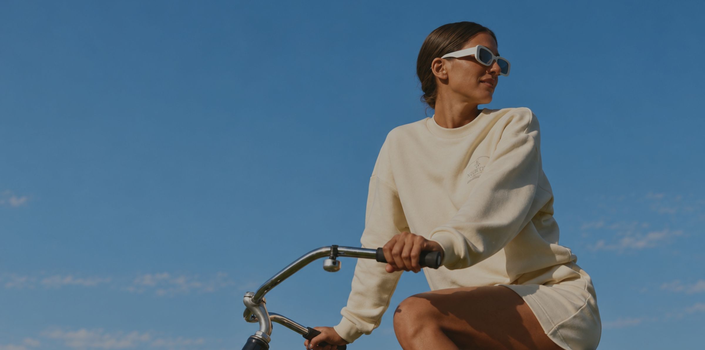 A woman in sunglasses and a cream sweatshirt riding a bicycle under a blue sky
