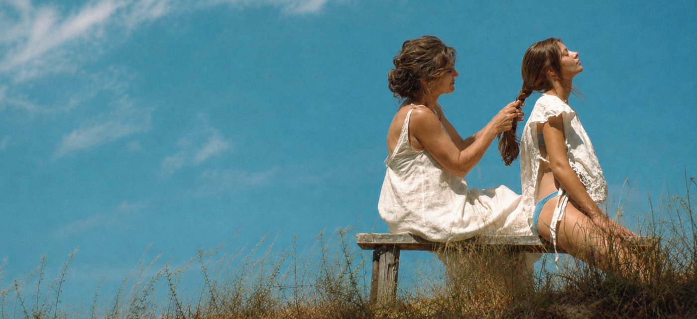 A woman braiding a younger woman's hair on a bench under a blue sky